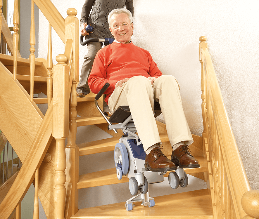 Das Bild zeigt einen älteren Herrn, der im Treppensteiger sitzt und eine Frau, die den Treppensteiger bedient. Man sieht den Steigvorgang der Treppenhilfe s-max sella. Der Steigfuß ist ausgefahren und setzt bereits auf der unteren Treppenstufe auf. Die Sicherheitsbremsen, welche am großen Laufrad befestigt sind, setzen am Stufenrand auf und schützen so vor einem abwärtsrollen. 